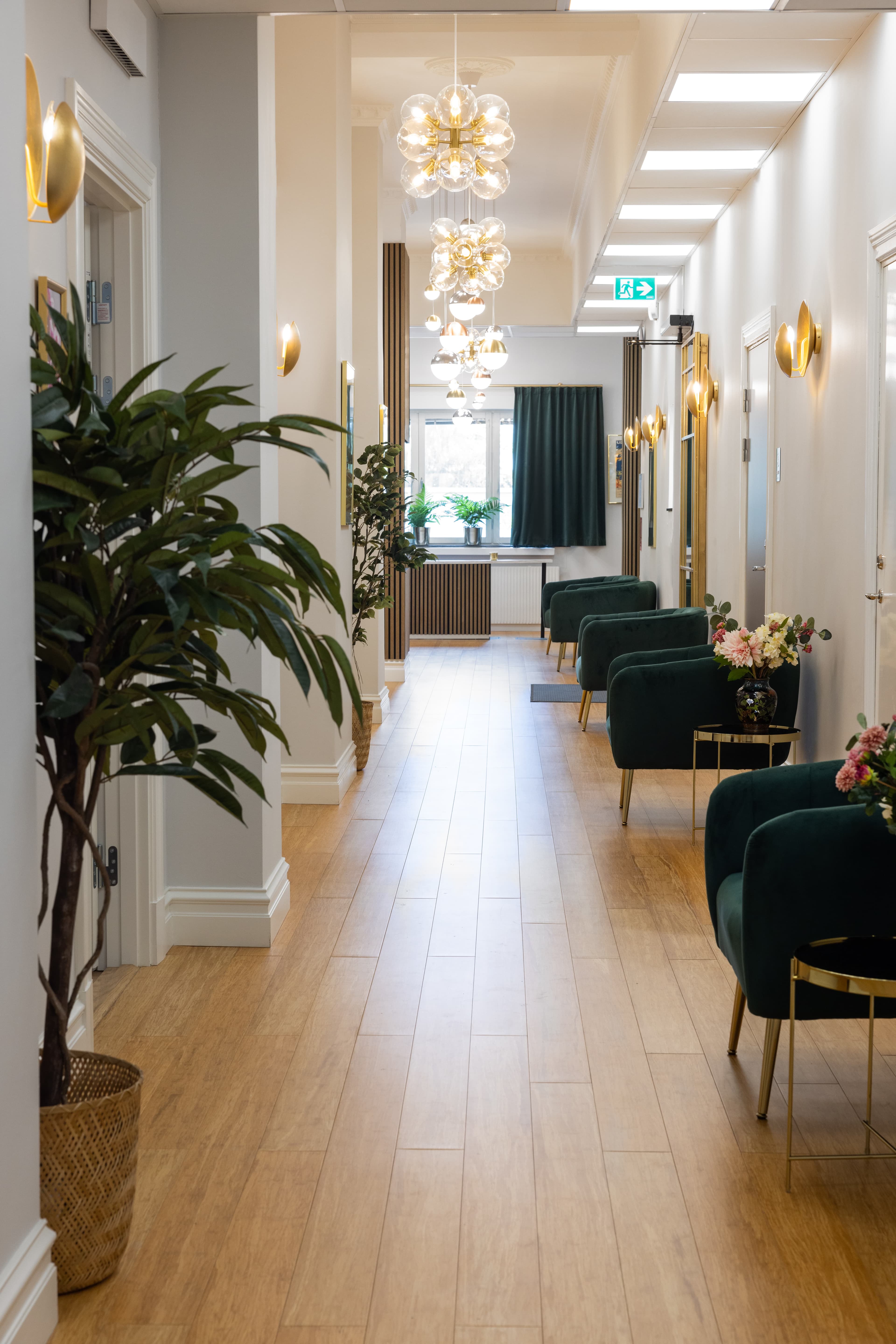 Elegant och inbjudande väntrum på en klinik för plastikkirurgi, med en stilren inredning i grönt och guld. Hallen är dekorerad med sammetsfåtöljer, små sidobord med blommor, moderna ljuskronor och varma vägglampor, vilket skapar en lyxig och professionell atmosfär.